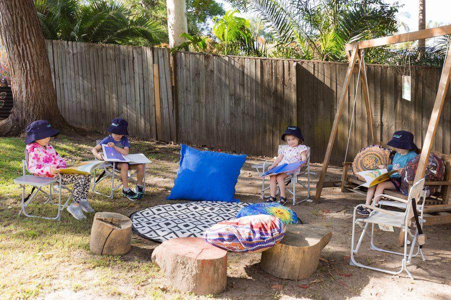 Children reading picture books on small chairs in the outdoor reading circle, with cushions and patterned mat