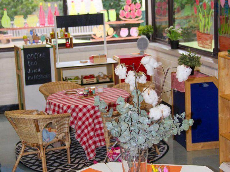 The home corner cafe area with cotton flowers in a vase, pencils in jars and play crockery on a red-checked table
