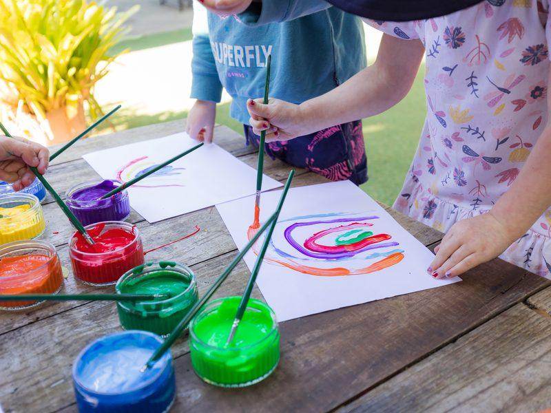 Children's rainbow paintings emerging on white paper, with paint pots and brushes on a wooden table